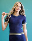 A woman modeling a navy blue textured knit short-sleeve top with gray and white trim, holding a hairbrush against a solid teal background.