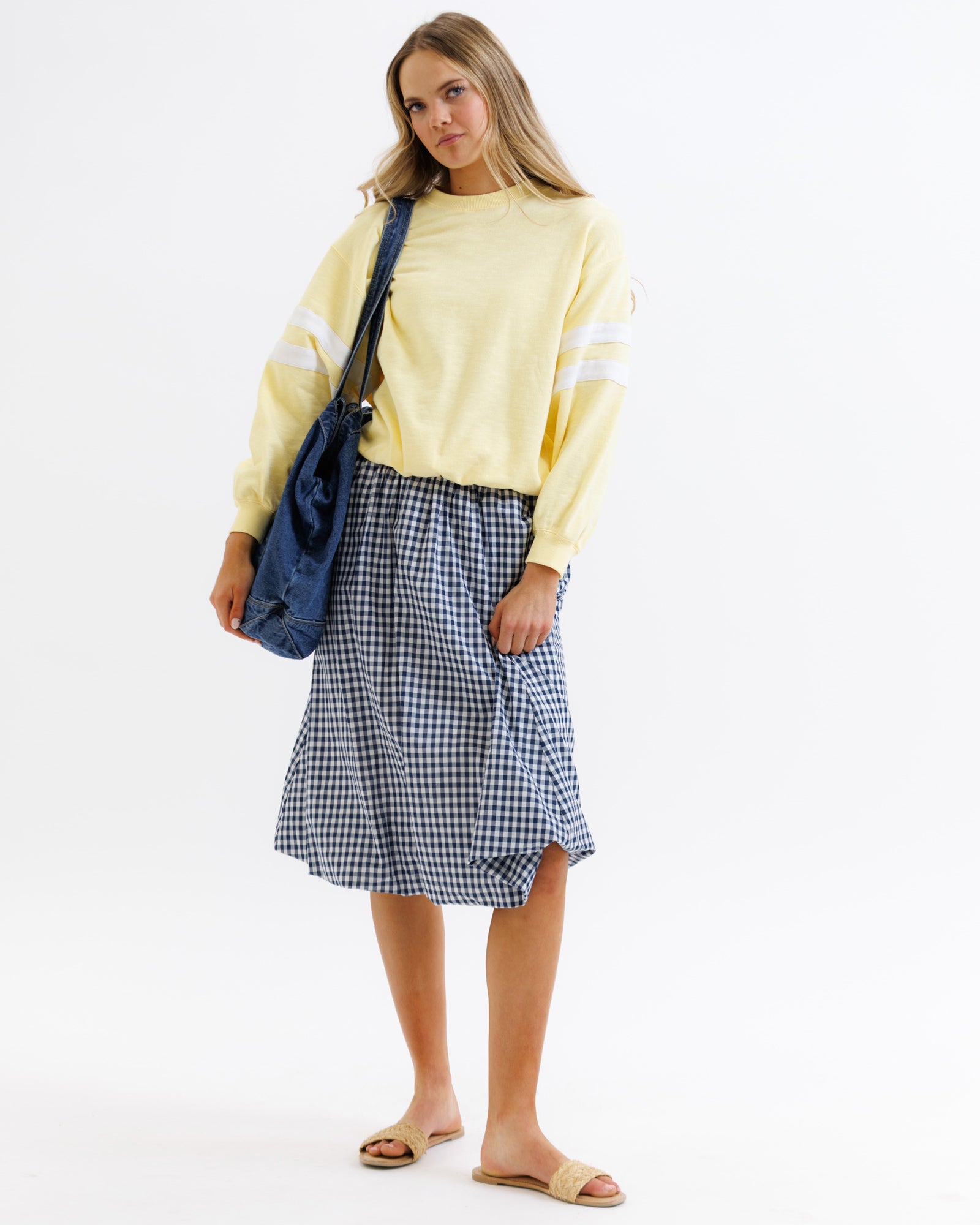 Woman wearing a yellow sweatshirt and blue checkered skirt with a blue bag on a white background
