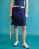 A person wearing a navy blue textured knit tennis skirt with a gray and white waistband, paired with white and navy Asics sneakers, holding a tennis racket against a teal background.