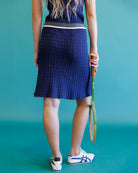 A woman from behind wearing a textured navy blue tennis skirt with a gray waistband, holding a tennis racket against a solid teal background.
