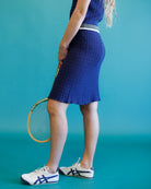 A woman wearing a textured royal blue knit tennis dress and white Asics sneakers, holding a tennis racket against a solid teal background.