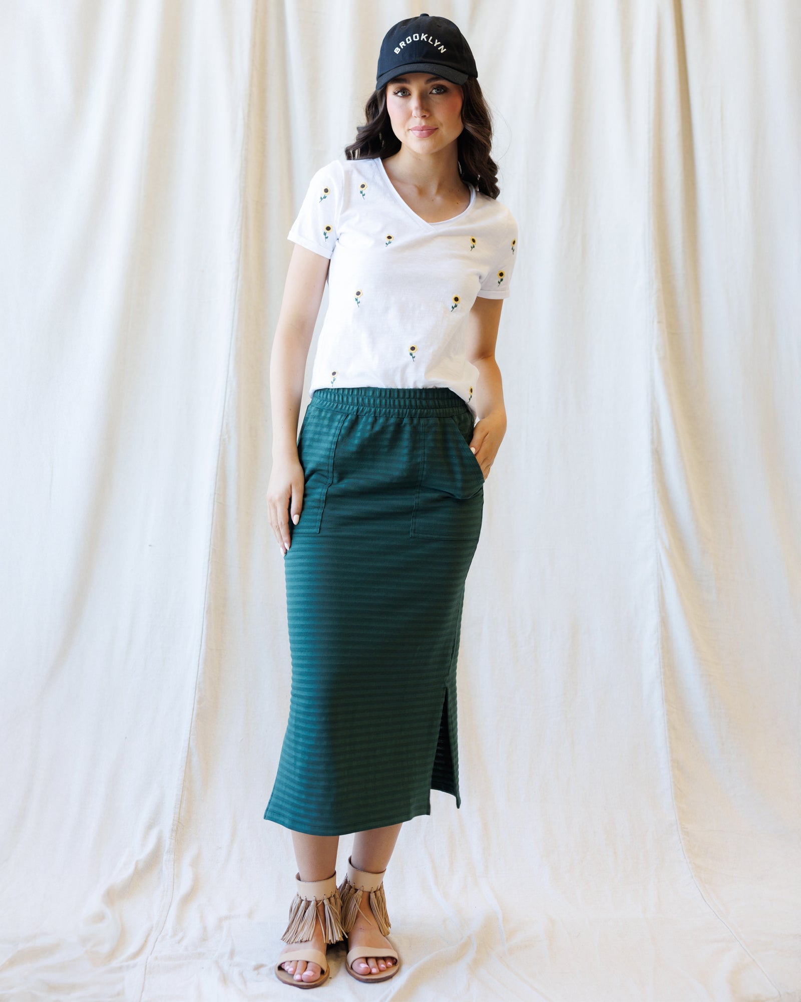 A woman modeling a dark green skirt with a white top, styled with tan strappy sandals against a draped white fabric backdrop.