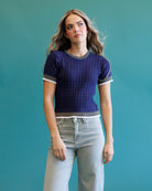 A woman modeling a short-sleeve textured knit top in deep purple with gray and white trim, paired with light-wash jeans, set against a solid teal background.