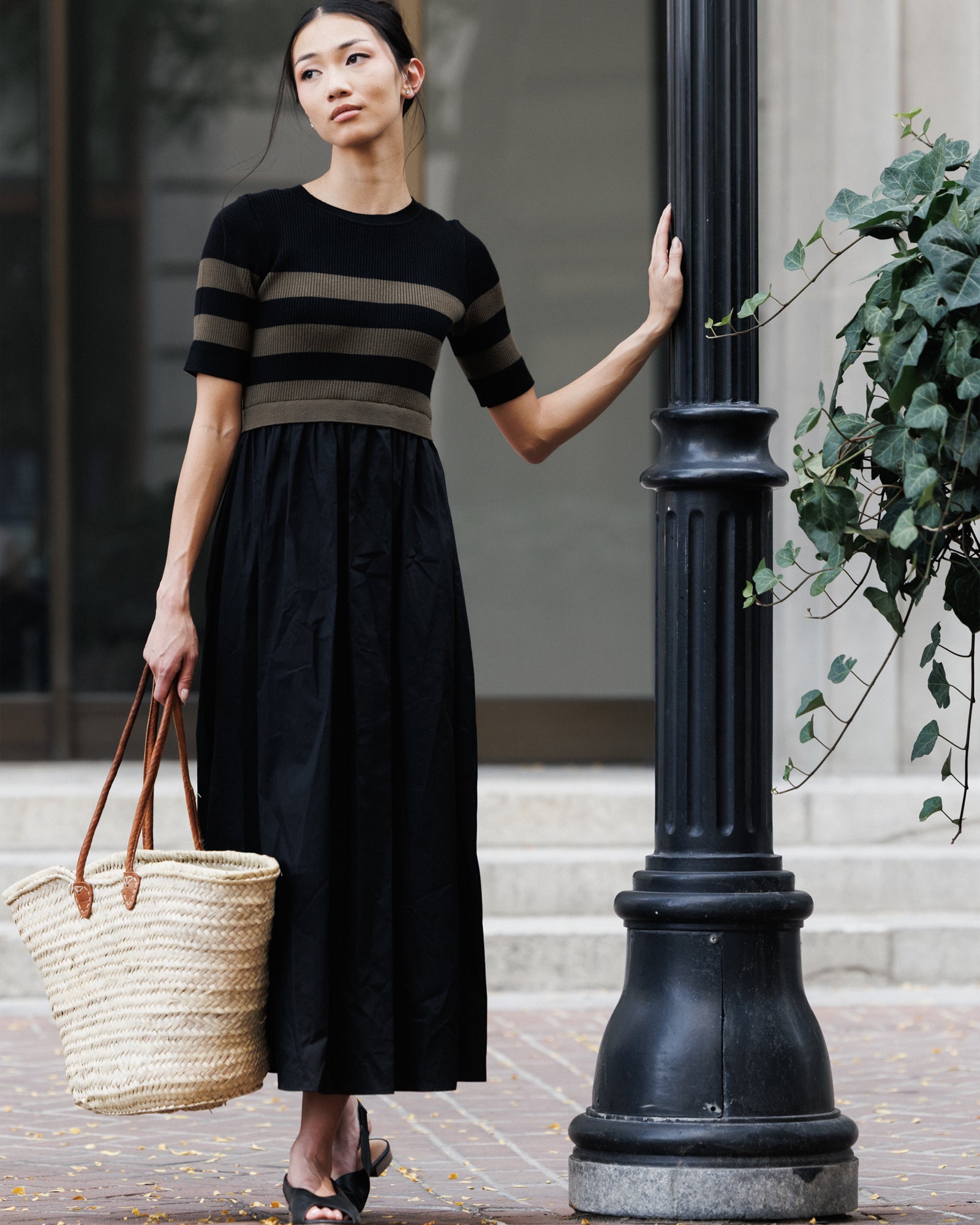 A woman stands on a city sidewalk beside a black lamppost, wearing a black midi dress with horizontal olive green stripes on the short sleeves and bodice. She holds a large woven straw tote bag with brown handles.