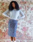 A woman stands with hands on hips wearing a light gray knit sweater with a white collar and a blue sequined skirt, posed in front of a vintage floral tapestry.