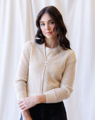 A woman wearing a cream-colored open-knit zip-up cardigan over a white top and black pants, standing against a soft white curtain background.