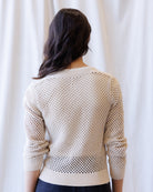 A woman viewed from behind wearing a cream-colored open-knit sweater with ribbed cuffs and hem, set against a soft white curtain background.