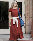 A woman wearing a rust-colored midi dress with a cream knit belt and carrying a blue denim shoulder bag, standing in front of a building entrance.
