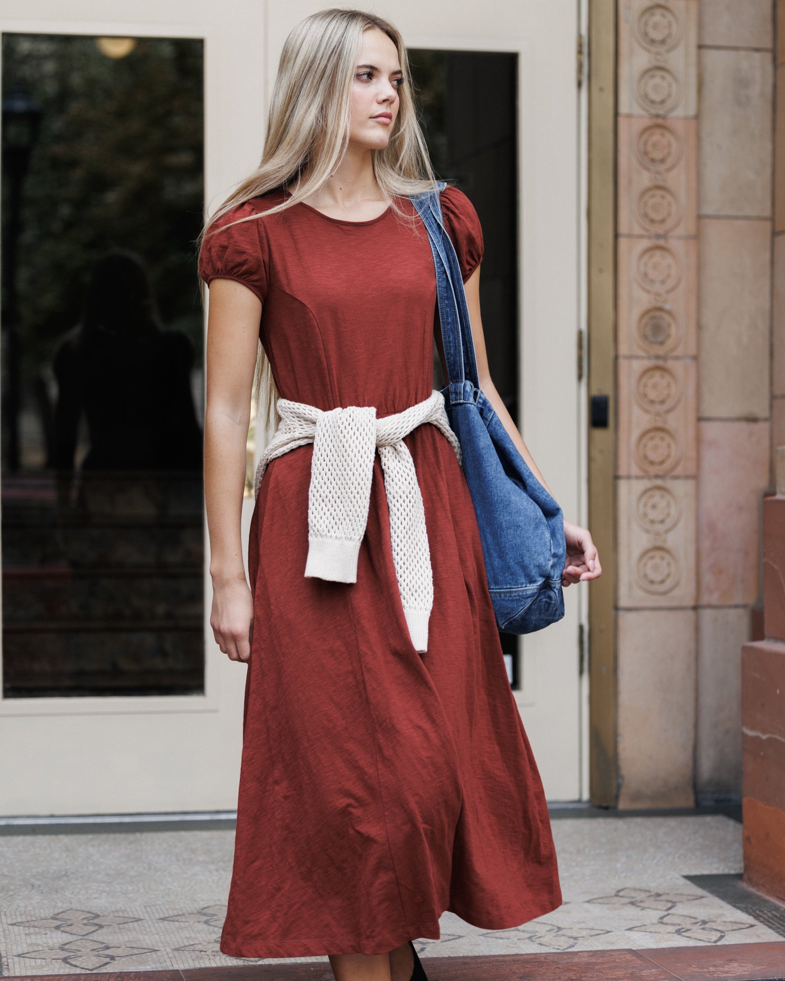 A woman wearing a rust-colored midi dress with a cream knit belt and carrying a blue denim shoulder bag, standing in front of a building entrance.