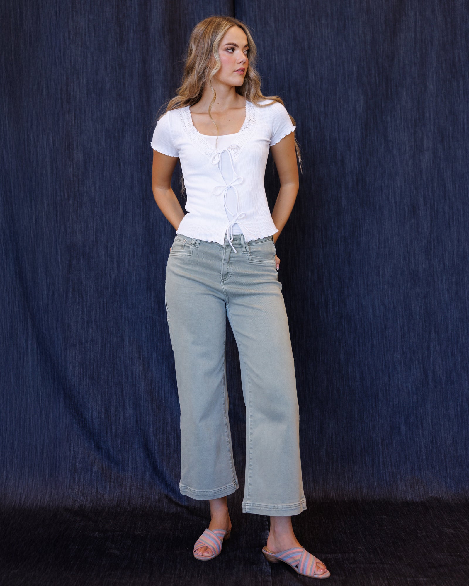 A woman modeling a white short-sleeve top with a square neckline and front tie detail, paired with light-wash wide-leg jeans and pink slide sandals, standing against a dark blue textured backdrop.