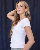 A woman with long wavy blonde hair wearing a white short-sleeve ribbed knit top with lace-up front details and scalloped edges, posed against a dark blue textured backdrop.