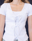 A woman wearing a white short-sleeve ribbed knit top with lace-up front details and scalloped edges, posed against a dark blue textured backdrop.