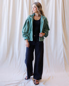 A woman modeling a sage green corduroy jacket over a black top and black wide-leg pants, standing against a draped cream fabric backdrop.