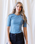 A woman with blonde wavy hair wearing a fitted dark blue short-sleeve crewneck top and dark jeans, standing against a draped white fabric background.