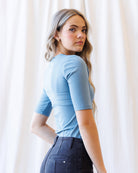 A woman with blonde wavy hair wearing a fitted dark blue short-sleeve crewneck top and dark jeans, standing against a draped white fabric background.