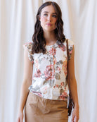 A woman wearing a sleeveless cream blouse with a pink and green floral print, paired with brown pants, standing against a soft white curtain backdrop.