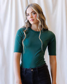A woman with blonde wavy hair wearing a fitted dark green short-sleeve crewneck top and dark jeans, standing against a draped white fabric background.
