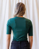 A woman with blonde wavy hair wearing a fitted dark green short-sleeve crewneck top and dark jeans, standing against a draped white fabric background.