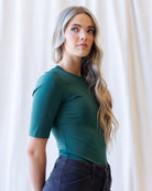 A woman with blonde wavy hair wearing a fitted dark green short-sleeve crewneck top and dark jeans, standing against a draped white fabric background.