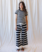 A woman modeling a matching black and white horizontally striped two-piece set, featuring a short-sleeve crewneck top and wide-leg pants with a white drawstring waist, standing against a draped white fabric backdrop.