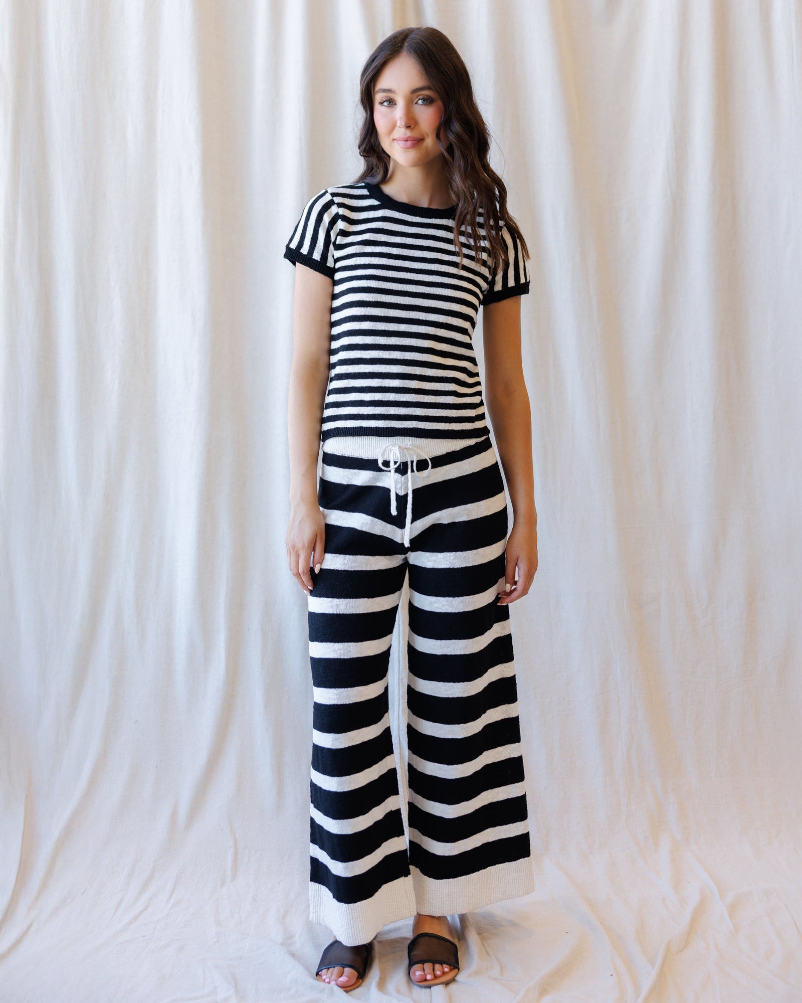A woman modeling a matching black and white horizontally striped two-piece set, featuring a short-sleeve crewneck top and wide-leg pants with a white drawstring waist, standing against a draped white fabric backdrop.