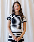 A woman wearing a short-sleeve knit top with horizontal black and white stripes, standing against a soft cream curtain backdrop.