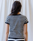 A woman viewed from behind wearing a short-sleeve knit top with horizontal black and white stripes, set against a soft cream-colored curtain background.