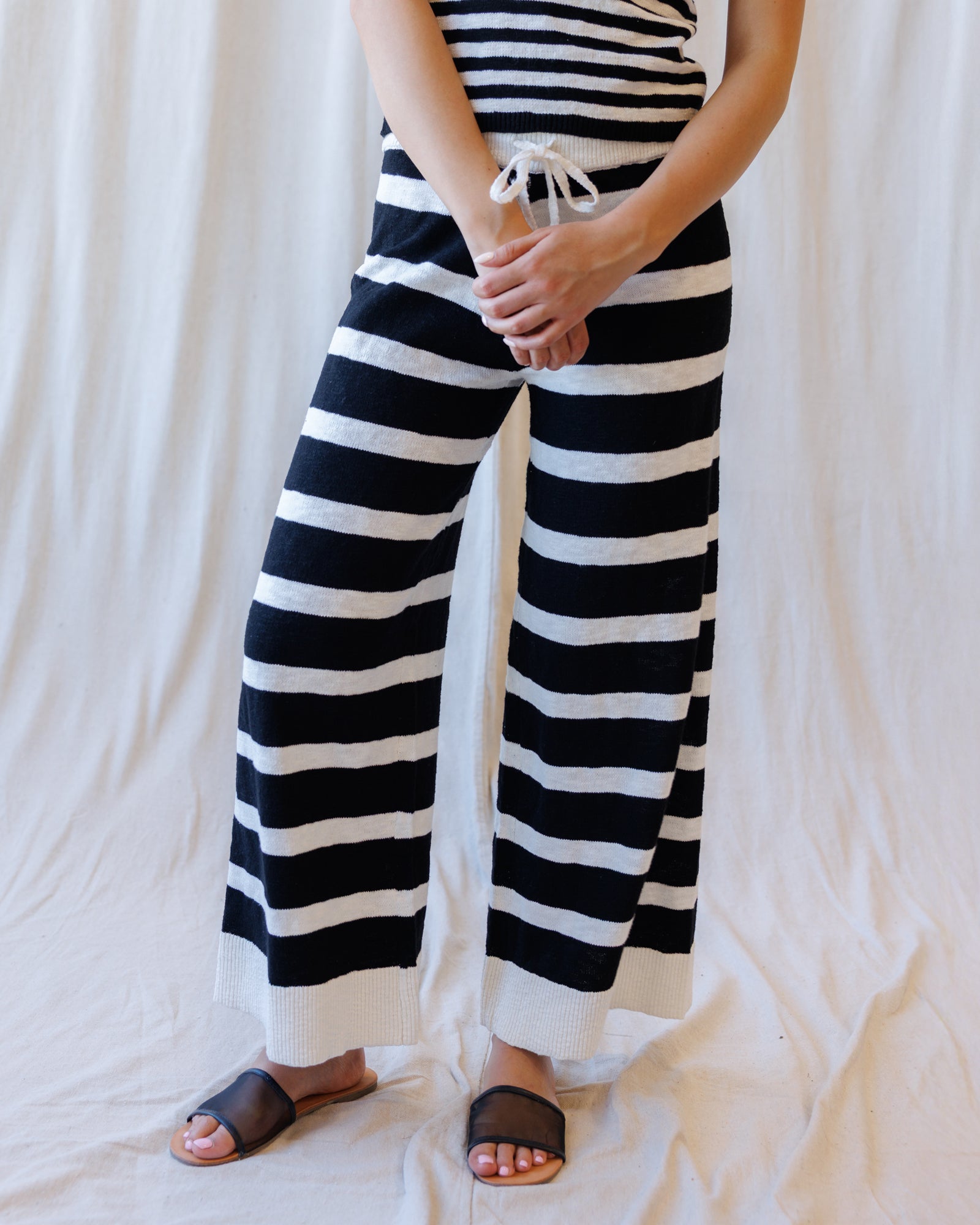 A person wearing black and white horizontally striped wide-leg knit pants with a drawstring waist, paired with dark brown slide sandals, standing against a draped white fabric backdrop.