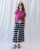 A woman modeling a magenta short-sleeve t-shirt and black and white horizontally striped wide-leg pants, styled with black sandals and sunglasses, standing against a draped white fabric backdrop.
