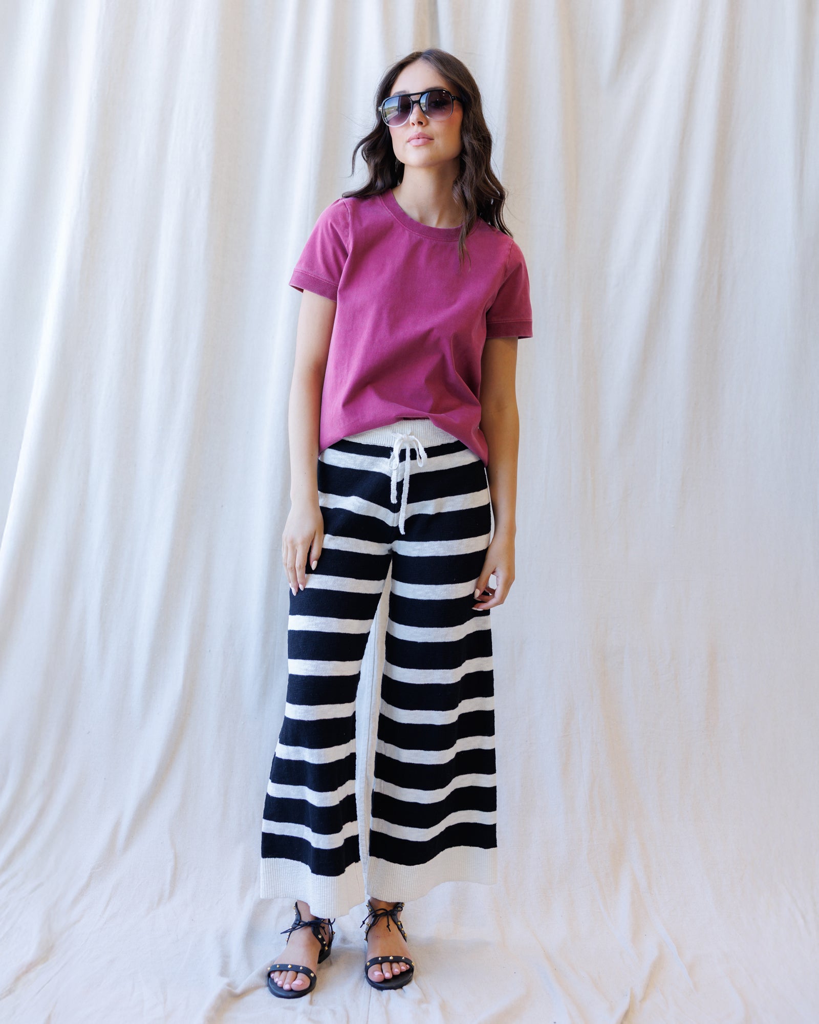 A woman modeling a magenta short-sleeve t-shirt and black and white horizontally striped wide-leg pants, styled with black sandals and sunglasses, standing against a draped white fabric backdrop.