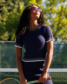 A woman on a tennis court wearing a navy blue textured short-sleeve dress with white and olive green trim, holding a tennis racket. The background shows a green tennis net and blurred trees.