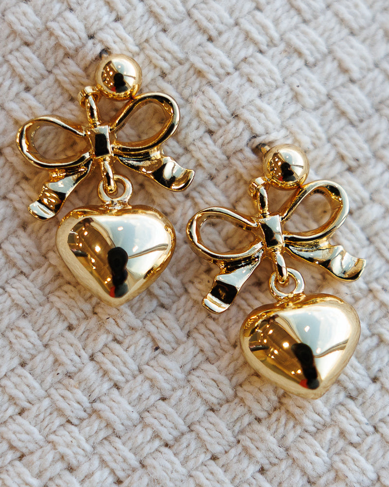 Gold bow and heart-shaped earrings on a textured beige surface