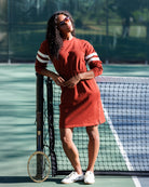 A woman stands on a tennis court wearing a rust-orange long-sleeve dress with white stripes on the sleeves, white sneakers, and sunglasses, leaning against a net with a tennis racket.