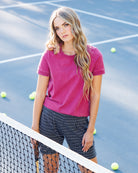 A woman stands on a blue tennis court wearing a short-sleeve crewneck t-shirt in magenta pink and black-and-white polka dot shorts, with tennis balls scattered around her and a net in the foreground.