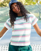 A woman on a tennis court wearing a white short-sleeve t-shirt with thin horizontal stripes in green, pink, and beige, paired with green shorts and a navy blue New York Yankees cap.
