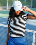 A woman on a tennis court wearing a white baseball cap with the word 'oui' in blue, a black and white horizontally striped knit top, and bright blue skirt or shorts.