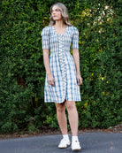 A woman stands outdoors in front of a green hedge wearing a short-sleeve blue and white plaid button-front dress with white sneakers.