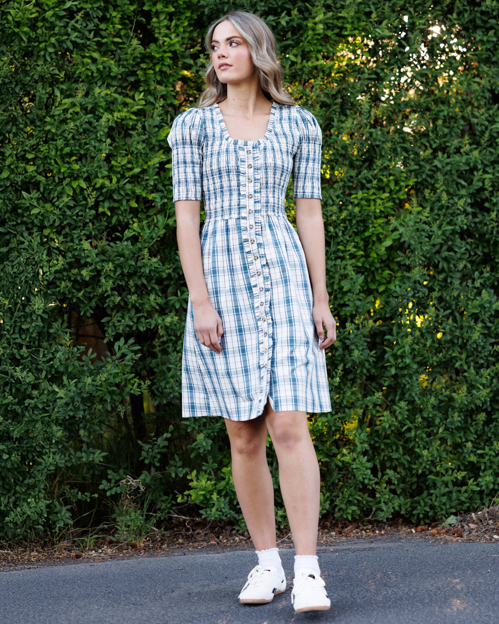 A woman stands outdoors in front of a green hedge wearing a short-sleeve blue and white plaid button-front dress with white sneakers.