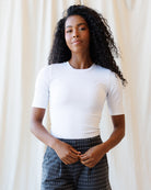 A woman with long, curly black hair is wearing a fitted white short-sleeve crewneck top and dark gray polka-dot pants, standing against a soft beige curtain backdrop.