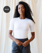 A  woman with long, curly black hair is wearing a fitted white short-sleeve crewneck top and dark gray polka-dot pants, standing against a soft beige curtain backdrop.