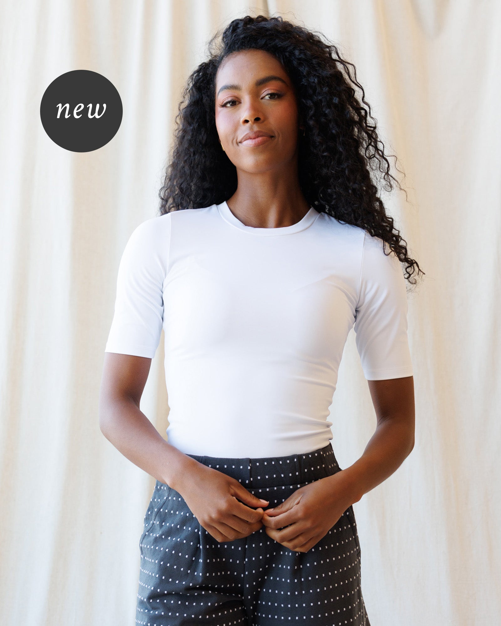 A  woman with long, curly black hair is wearing a fitted white short-sleeve crewneck top and dark gray polka-dot pants, standing against a soft beige curtain backdrop.
