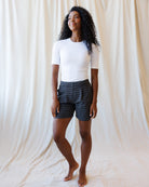 A woman with curly black hair stands barefoot in front of a draped cream fabric backdrop, wearing a fitted white short-sleeve top and dark shorts with a small white geometric pattern.