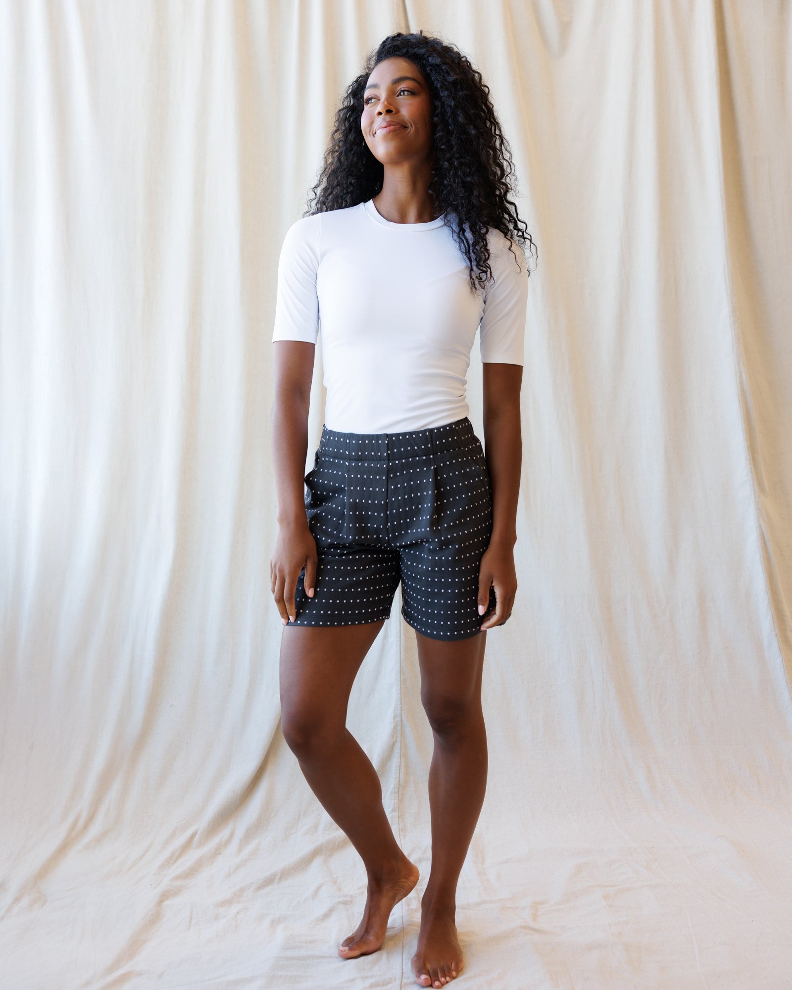 A woman with curly black hair stands barefoot in front of a draped cream fabric backdrop, wearing a fitted white short-sleeve top and dark shorts with a small white geometric pattern.