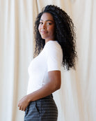A woman with long, curly black hair is shown in profile, wearing a fitted white short-sleeve top and dark gray pants with small white polka dots, standing against a draped cream fabric backdrop.