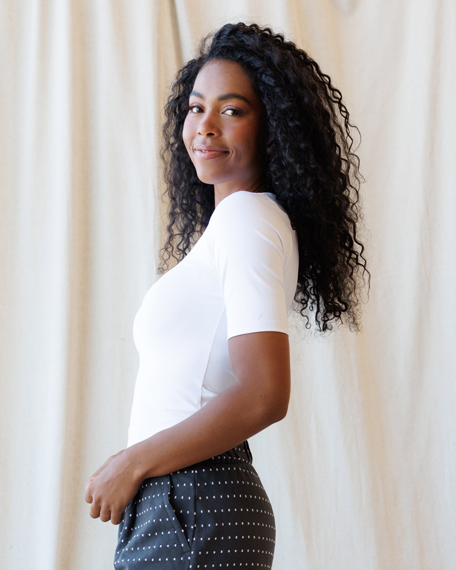 A woman with long, curly black hair is shown in profile, wearing a fitted white short-sleeve top and dark gray pants with small white polka dots, standing against a draped cream fabric backdrop.