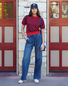 Person wearing a checkered shirt, jeans, and a cap standing in front of a building with red doors.