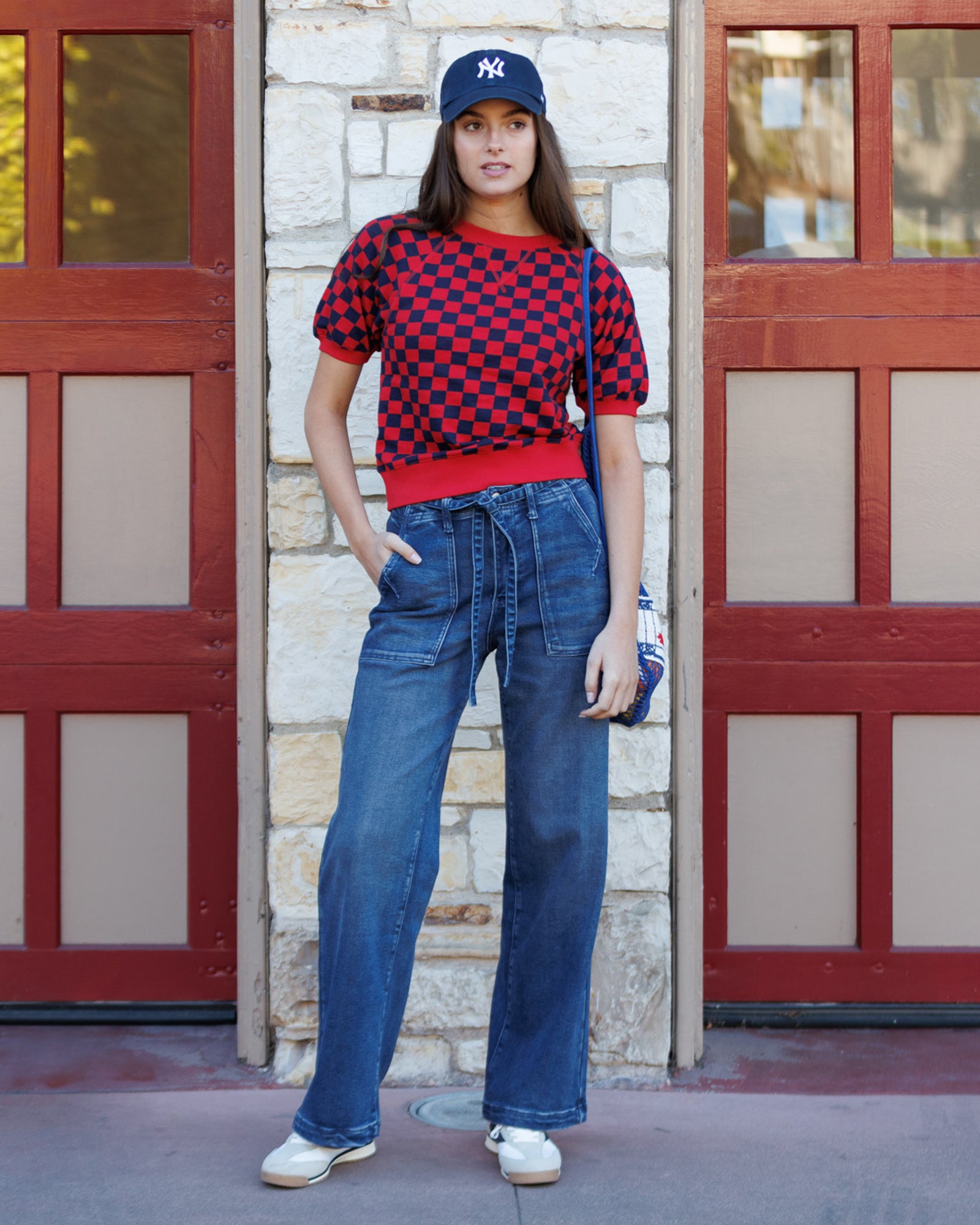 Person wearing a checkered shirt, jeans, and a cap standing in front of a building with red doors.