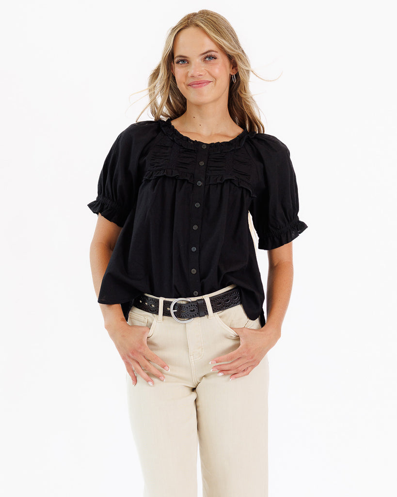 Woman wearing a black blouse with ruffled details and beige pants on a white background