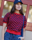 Woman wearing a red and blue checkered sweater with a blue cap and jeans, standing against a blurred background.