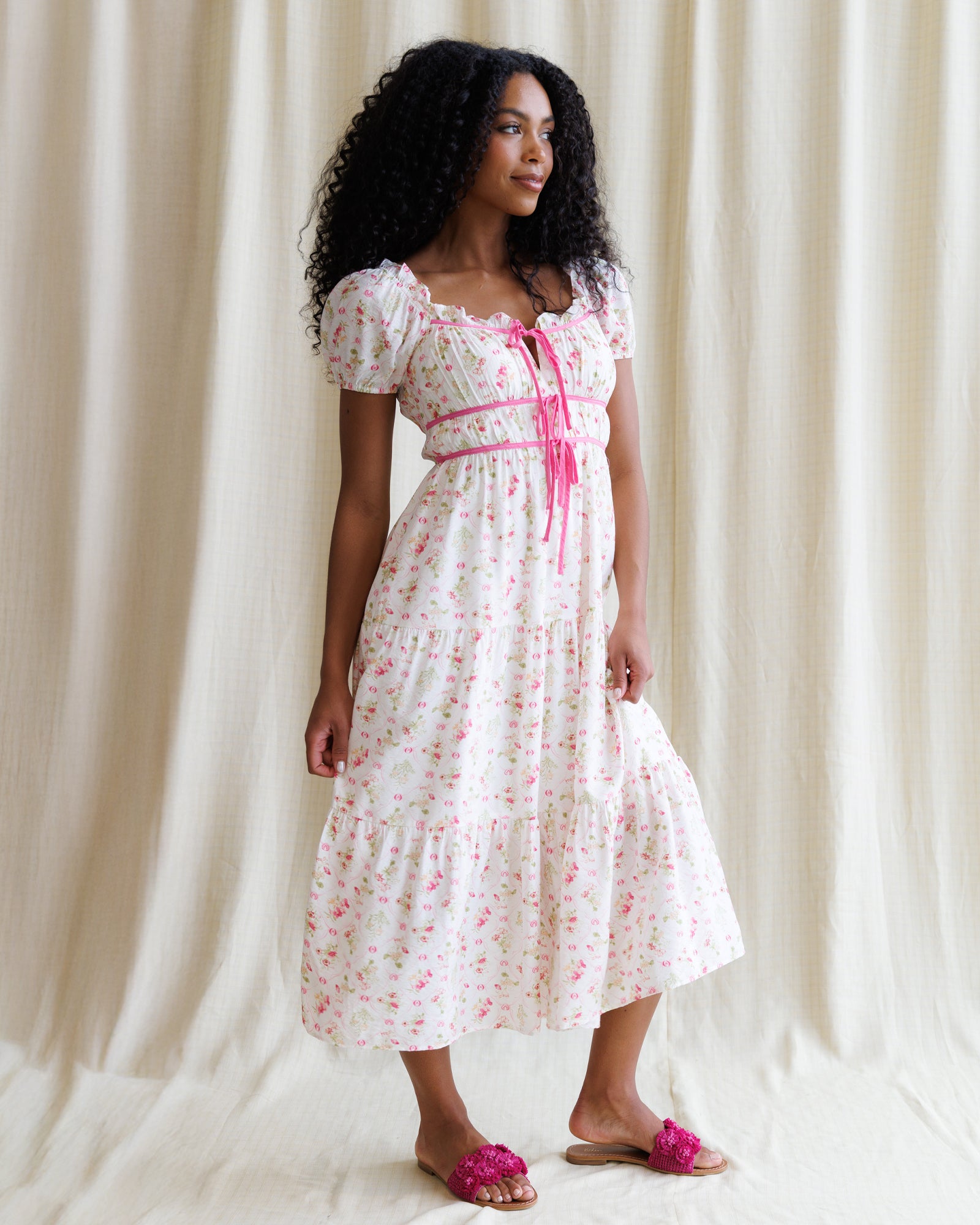 Woman wearing a floral dress with a pink belt against a white curtain background